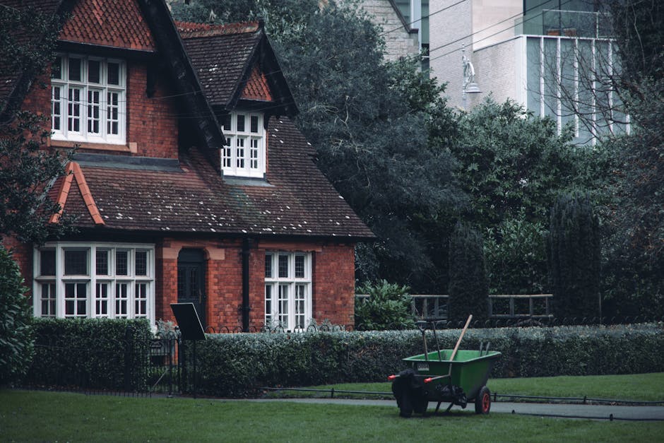 A small, neatly maintained front garden of a red-brick house with a sloped, tiled roof and white-framed windows, featuring detailed latticework on the upper windows and a dark entrance door. The garde